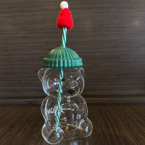 Clear bear-shaped glass with a green ridged lid, a striped straw, and a red knitted hat.