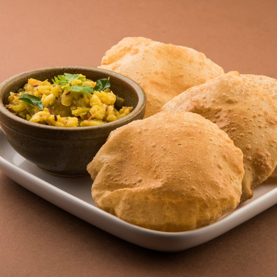 A bowl of yellow potato curry with cilantro and cumin seeds, served with three golden-brown puri breads.